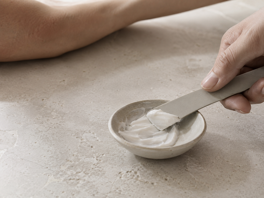 Main prélevant une crème dépilatoire blanche avec une spatule sur une surface minérale claire.